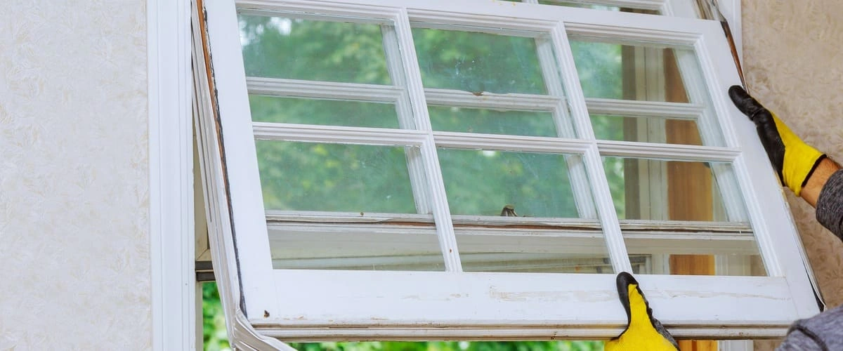 A close-up view of a technician wearing yellow work gloves lifting an old white wooden sash during a home window replacement project.