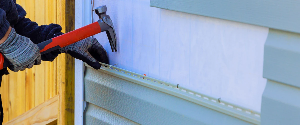 Close up view of a worker wearing gloves using a red hammer to secure light green vinyl siding panels to an exterior home wall during a renovation.