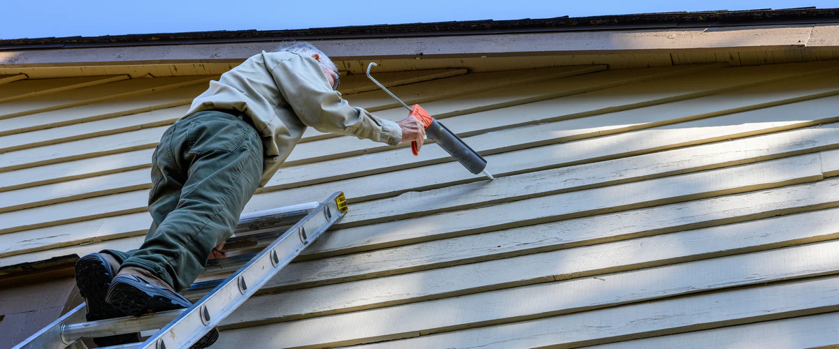 A professional contractor standing on a ladder while using a caulking gun to apply waterproof sealant to the gaps in horizontal wooden house siding.