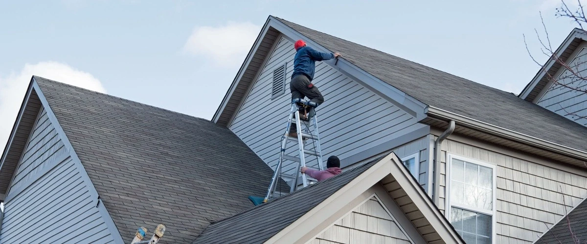 Two workers performing home repairs on a large multi-story house, with one technician on a tall ladder inspecting the grey vinyl siding and roof trim against a clear sky.