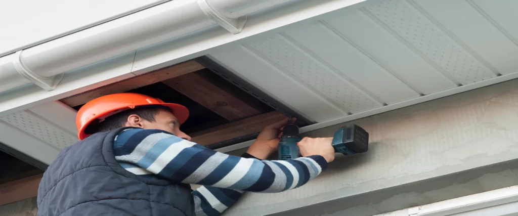 A professional contractor in an orange hard hat using a power drill to install white vented soffit panels under a roof eave.