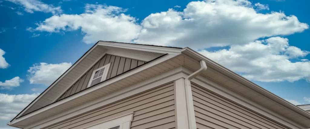 Low angle view of tan horizontal siding and white soffit under the roof eaves against a cloudy blue sky.