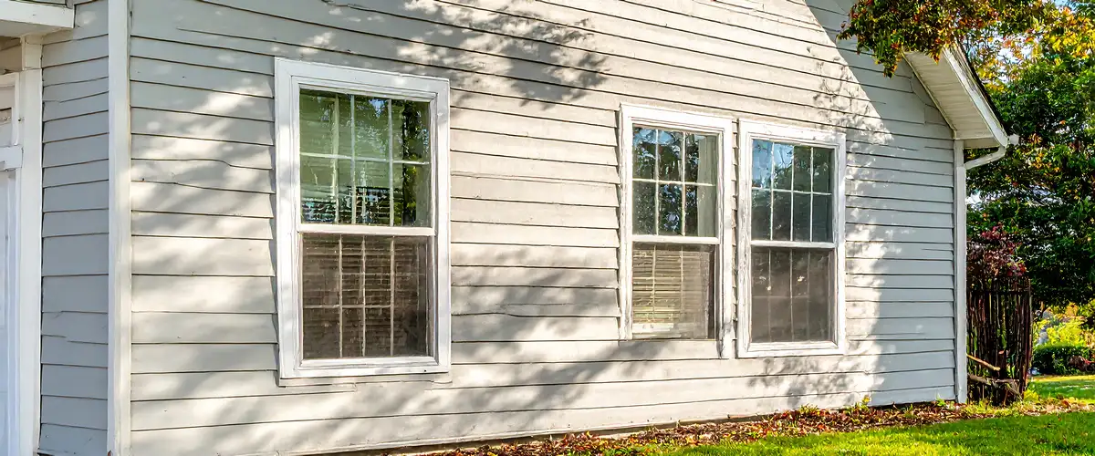 The exterior side wall of a house showing traditional gray wood lap siding in natural sunlight with three white-trimmed windows and shadows from nearby trees.