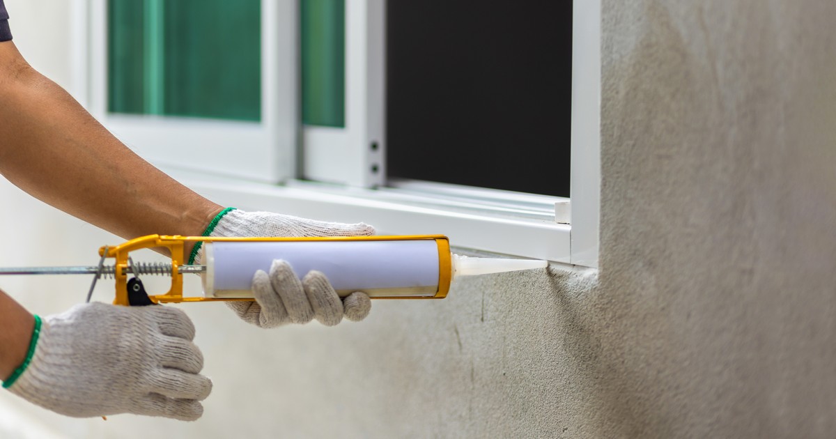 A worker wearing white gloves uses a yellow caulking gun to apply Adfast caulking for windows along the bottom track of a white sliding window against a concrete exterior wall.