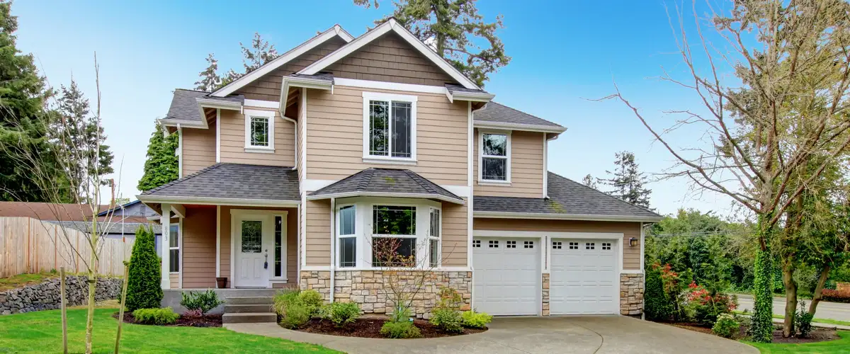 Large two-story residential home exterior showcasing tan horizontal siding paired with natural stone veneer accents and white garage doors.