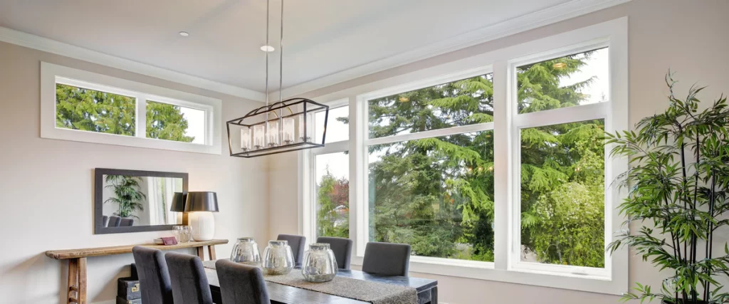 A dining room with a dark wooden table and modern chandelier, featuring a stunning full frame window installation that offers expansive views of tall green pine trees.