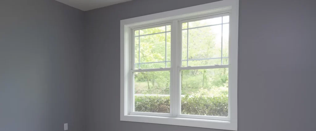 A single, double-hung pocket insert window with white trim and decorative perimeter grilles installed in a smoothly painted grey wall overlooking green foliage.