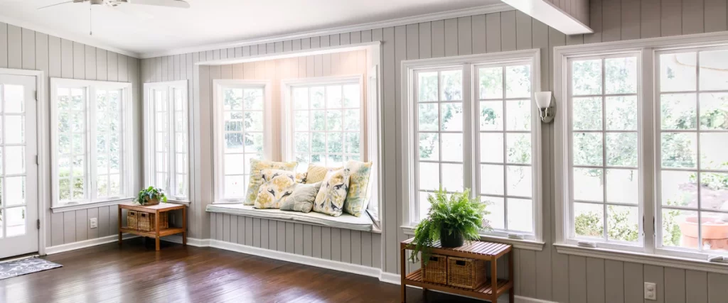 A cozy sunroom featuring a bay window seat with decorative pillows, surrounded by tall, multi-pane energy efficient windows that let in abundant natural light over a wood floor.