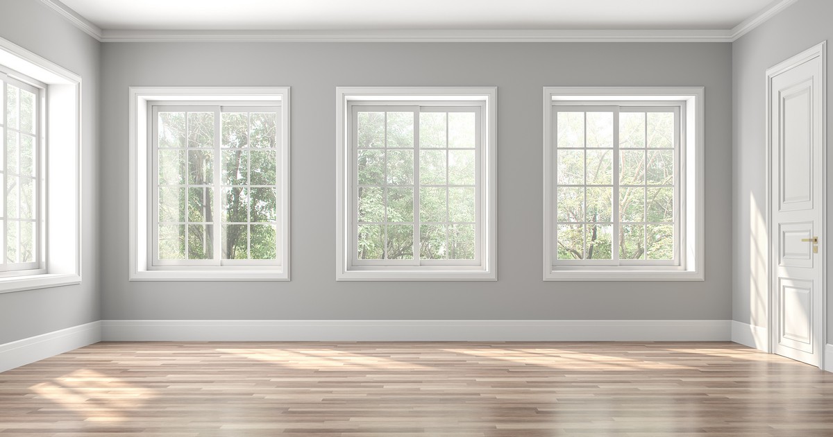 An empty room with light grey walls and hardwood floors showcasing a recent Full Frame Window Replacement with three large, white-framed windows letting in bright sunlight.