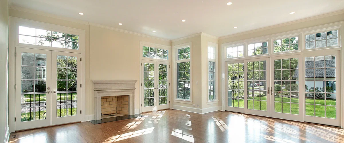 A sunlit, empty living room with hardwood floors featuring a large white-framed grid window and matching French doors next to a stone fireplace.