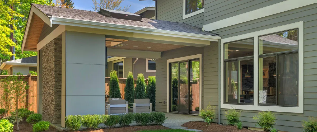 Modern sage green house exterior showing a large picture window and sliding glass door under a covered patio with wood-plank ceilings and stone accents.