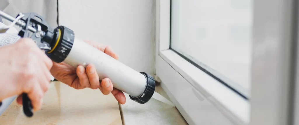 A close-up shot of a professional using a silver sausage caulking gun to apply a smooth bead of white sealant to the joint between a white window frame and a marble windowsill.