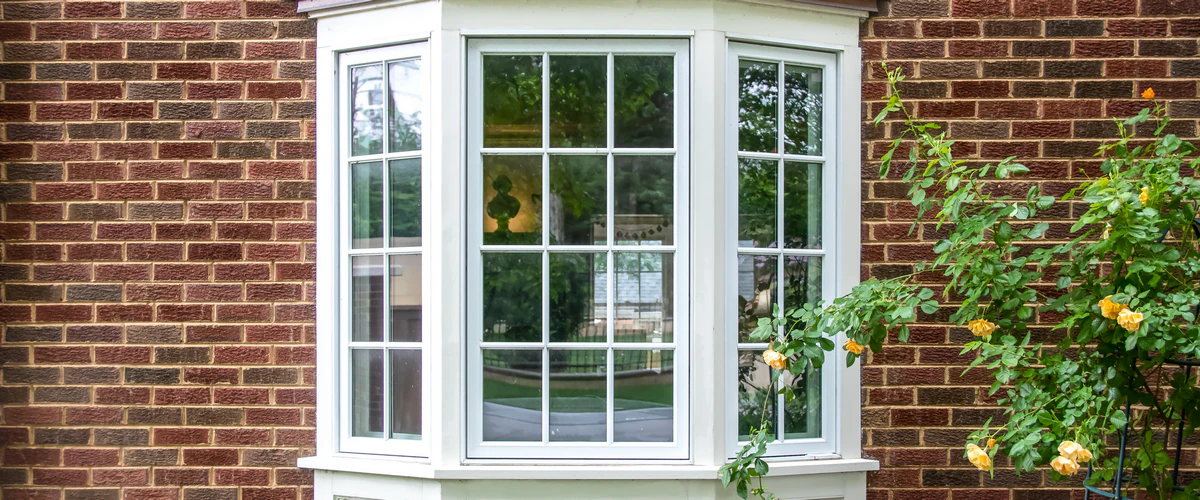 Elegant white bay window with decorative grids installed in a classic red brick wall with yellow roses growing on the side.