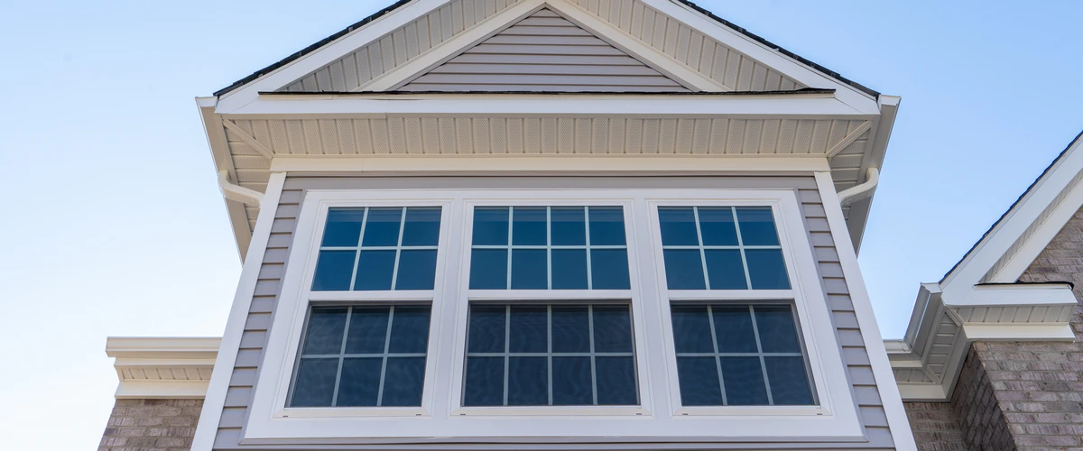 A close-up, low-angle shot of a white-trimmed window unit with grids installed on a grey-sided house under a gabled roofline.