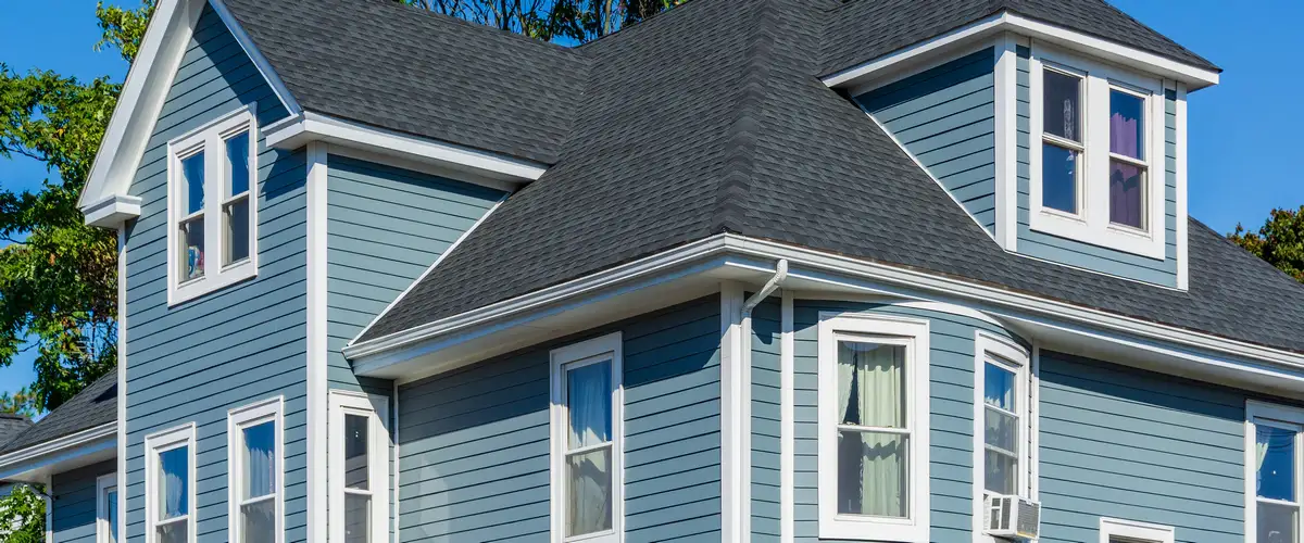 Two-story house exterior featuring blue horizontal siding, crisp white window trim, and a dark shingle roof under a clear sky.