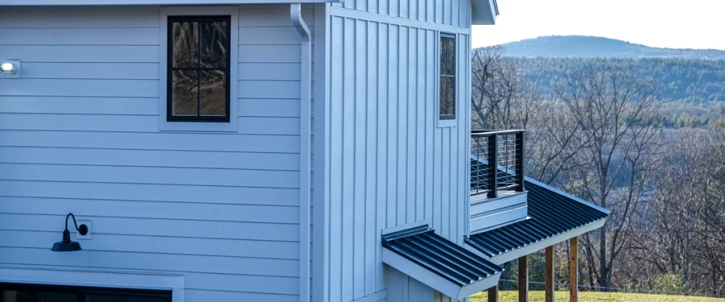 Close-up architectural detail of a house exterior showing crisp white James Hardie vertical and horizontal siding, a black metal window awning, second-story balcony railings, and dark gooseneck wall lights with a scenic hillside background.