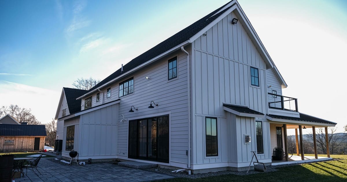 A wide exterior view of a large modern white farmhouse showing mixed vertical and horizontal siding, a stone patio, large black-framed windows, and durable James Hardie® Soffit under the roof eaves.