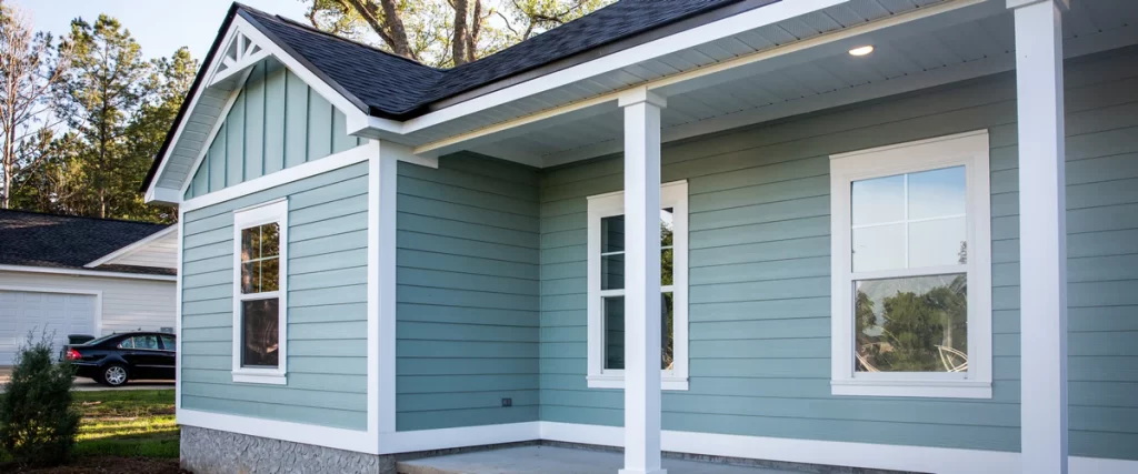 Single-story home with a front porch, white pillars, and light teal horizontal siding against a backdrop of trees and a blue sky.
