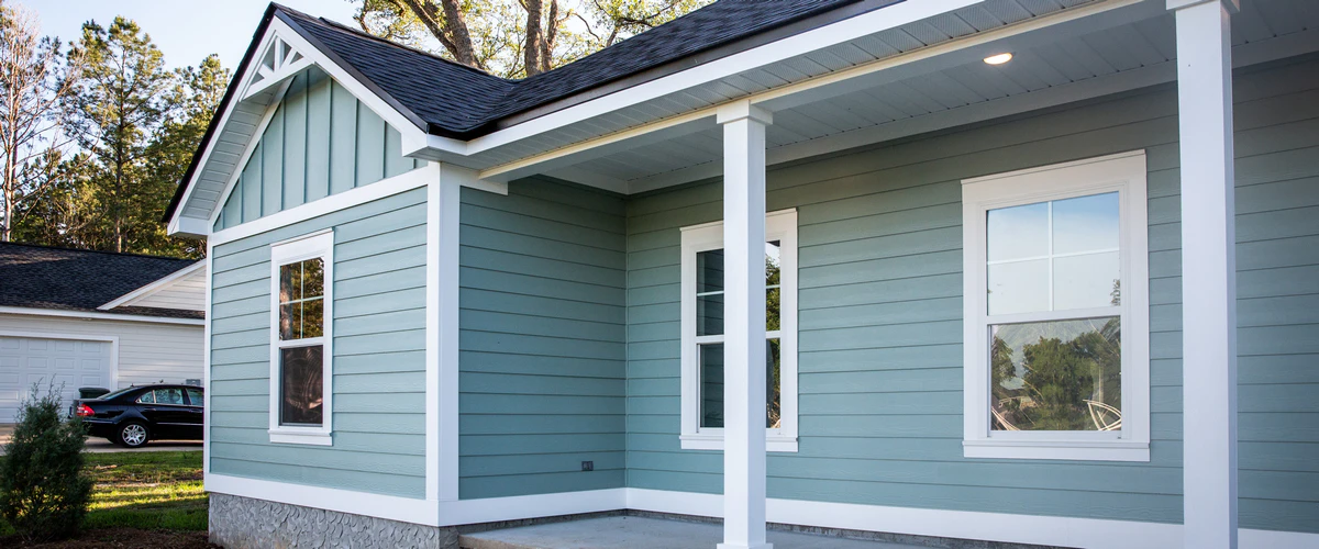 Single-story home with a front porch, white pillars, and light teal horizontal siding against a backdrop of trees and a blue sky.