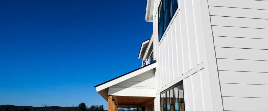 An angled view of a crisp white modern farmhouse exterior featuring James Hardie vertical board-and-batten alongside horizontal lap siding, black windows, and a wood-post covered porch against a clear blue sky.
