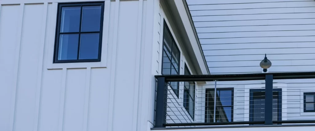 A modern white home exterior featuring vertical board-and-batten siding, a sleek black balcony railing with wire cables, and energy efficient dark-framed windows.